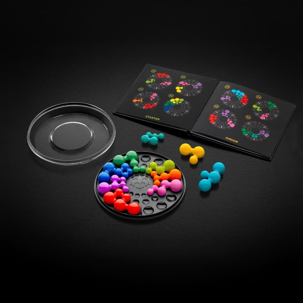 A colourful logic puzzle game set displayed on a dark background, including a clear round lid, a black circular tray filled with brightly coloured pieces, loose coloured pieces, and the challenge book