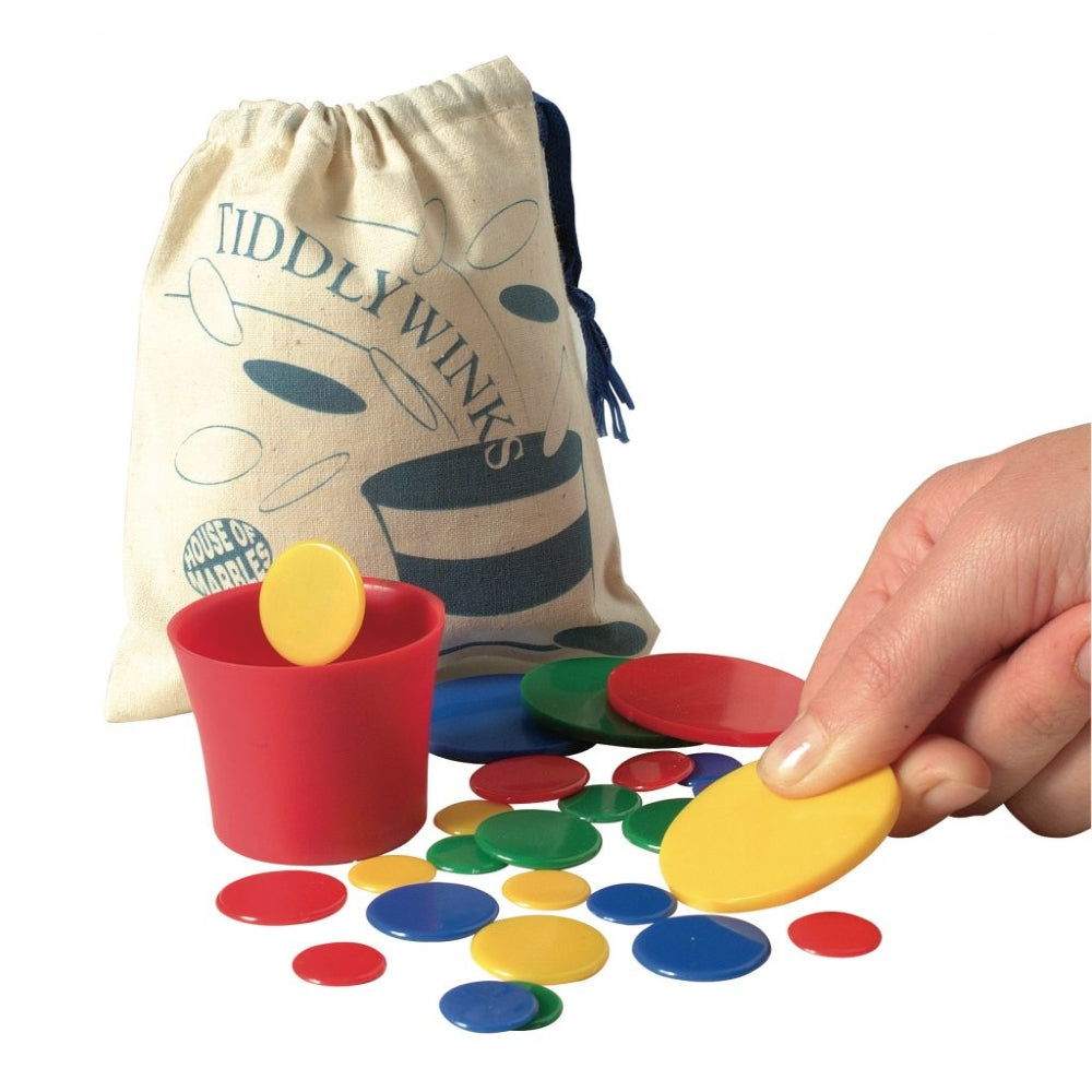 A hand holding a yellow counter next to a red cup with a yellow counter falling in, with scattered red, blue, green, and yellow counters in front. A drawstring pouch printed with 'Tiddlywinks' stands behind the game on a white background.